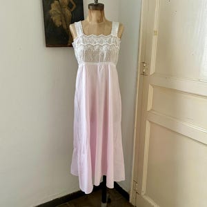 May include: A sleeveless nightdress featuring a light pink skirt and a white bodice with intricate eyelet lace embroidery. The bodice has a square neckline with a scalloped edge and wide straps, transitioning into a simple, flowing light pink skirt.