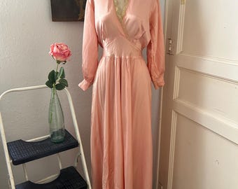 Könnte beinhalten: Ein vintage pfirsichfarbenes Kleid mit langen Ärmeln und einem Wickel-Oberteil wird auf einer Schaufensterpuppe präsentiert. Das Kleid hat einen fließenden Rock und eine zarte goldene Verzierung am Ausschnitt. Das Kleid ist aus einem seidigen Material gefertigt und hat ein klassisches, elegantes Design.