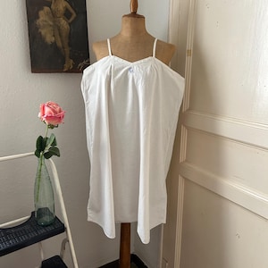 Puede incluir: Un camisón blanco con tirantes finos se exhibe en un maniquí de madera. El camisón tiene un escote en V y un pequeño detalle bordado en azul. El maniquí está colocado contra una pared con un póster vintage y una rosa rosa en un jarrón.