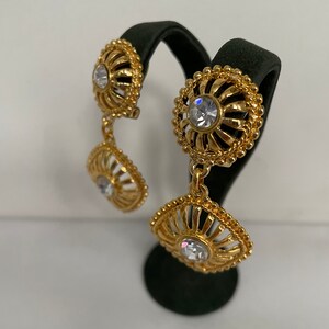 May include: Gold tone dangle earrings with clear crystal accents. The earrings feature a delicate, ornate design with a circular and square shape.