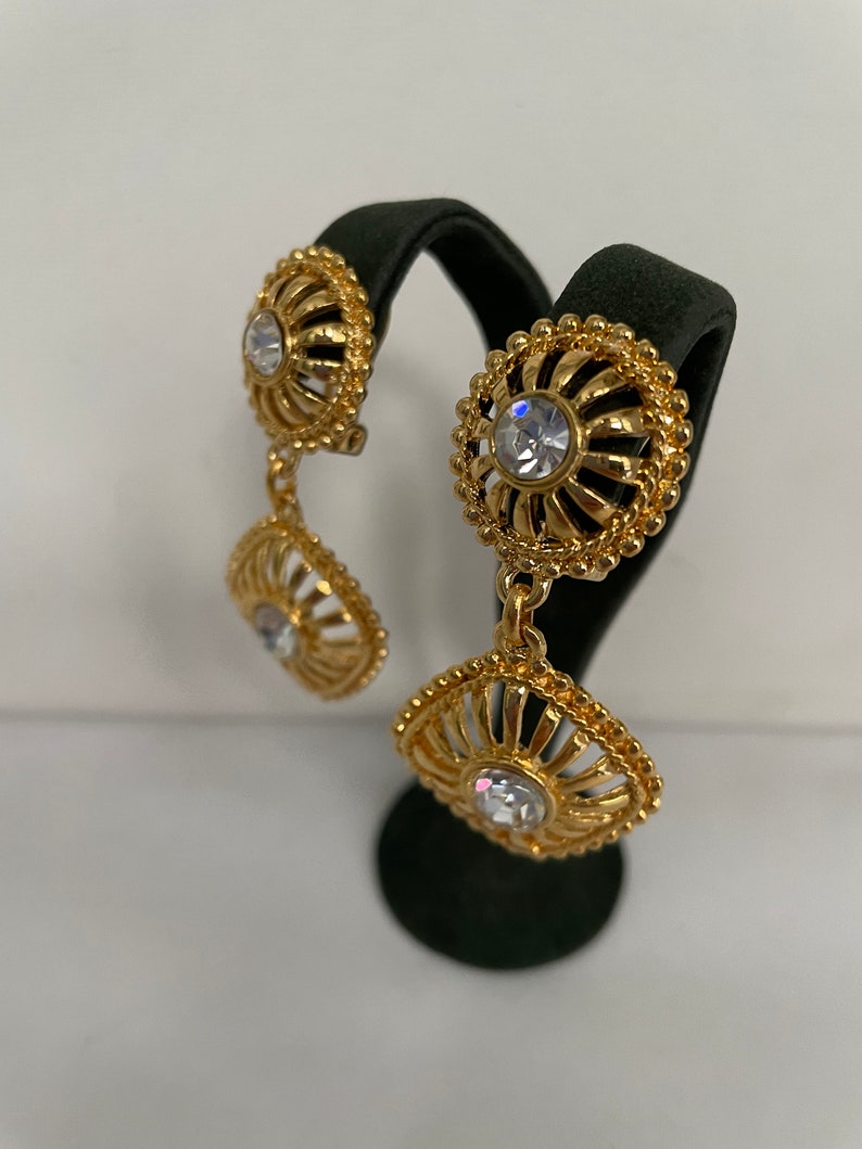 May include: Gold tone dangle earrings with clear crystal accents. The earrings feature a delicate, ornate design with a circular and square shape.