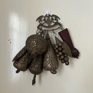 Puede incluir: Un adorno de pared de latón con una pieza superior arqueada y ornamentada con motivos de aves. De ella cuelgan varios dijes de latón texturizado, incluyendo adornos esféricos y en forma de pera, un racimo de uvas y una figura de un brazo.