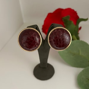May include: A pair of round, burgundy earrings with a gold rim. The earrings have a textured, reptile-like pattern.