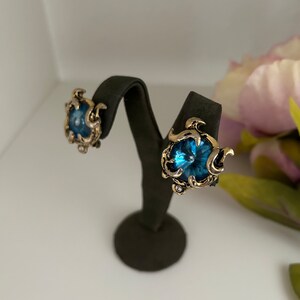 K&ouml;nnte beinhalten: Ein Paar Ohrclips im Vintage-Stil. Jeder Ohrring hat einen gro&szlig;en, runden, blauen Edelstein, der in einen goldfarbenen, verzierten Rahmen gefasst ist. Die Ohrringe werden auf einem schwarzen Ohrringst&auml;nder vor wei&szlig;em Hintergrund pr&auml;sentiert.