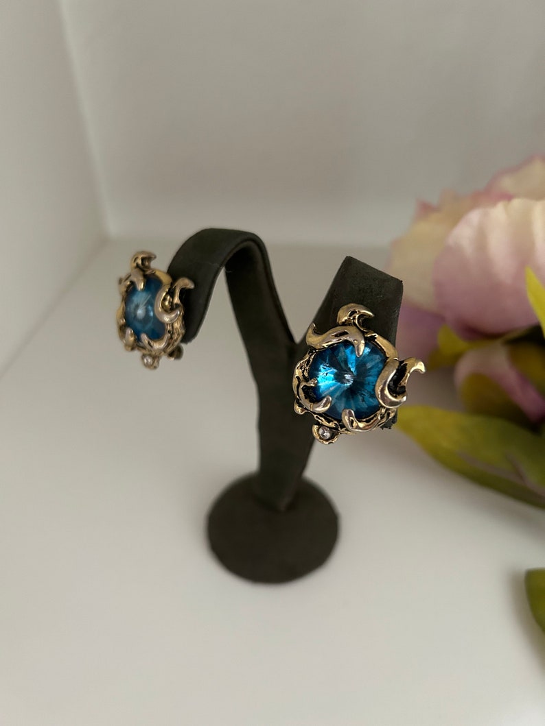 K&ouml;nnte beinhalten: Ein Paar Ohrclips im Vintage-Stil. Jeder Ohrring hat einen gro&szlig;en, runden, blauen Edelstein, der in einen goldfarbenen, verzierten Rahmen gefasst ist. Die Ohrringe werden auf einem schwarzen Ohrringst&auml;nder vor wei&szlig;em Hintergrund pr&auml;sentiert.