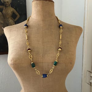 May include: A gold chain necklace with blue, green, and red oval-shaped beads. The necklace is on a mannequin.