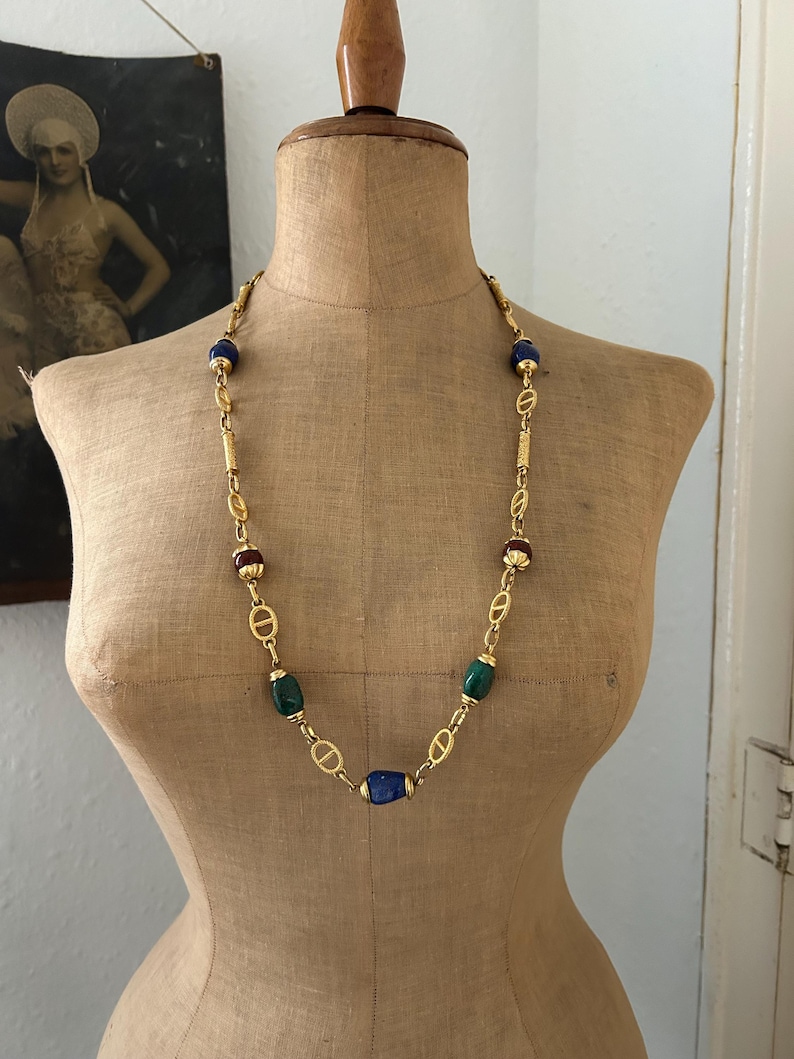May include: A gold chain necklace with blue, green, and red oval-shaped beads. The necklace is on a mannequin.