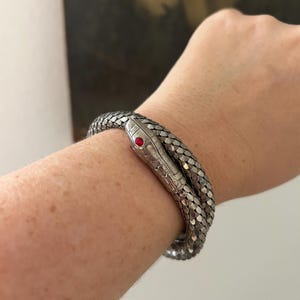 May include: Silver-toned snake bracelet with a red gemstone eye. The bracelet is wrapped around a wrist, showcasing the intricate scale-like design. The snake's head is detailed, adding a realistic touch to the jewelry.