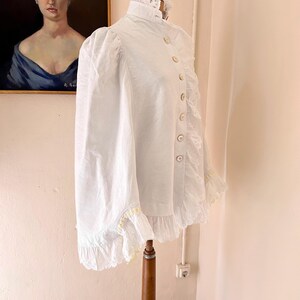 May include: A white Victorian-style blouse with a high collar and long sleeves. The blouse features decorative buttons down the front and lace trim along the collar, cuffs, and hem. The garment is displayed on a wooden dress form.