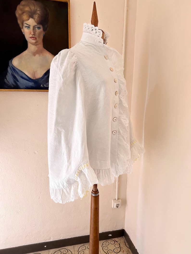 May include: A white Victorian-style blouse with a high collar and long sleeves. The blouse features decorative buttons down the front and lace trim along the collar, cuffs, and hem. The garment is displayed on a wooden dress form.