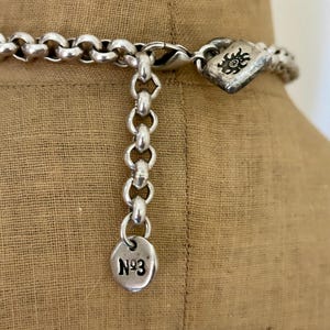 May include: Close-up of a silver-toned necklace with a large link chain. The necklace has a decorative clasp with a sunburst design and a small circular charm with the number "N&ordm;3" engraved on it. The necklace is displayed on a tan fabric.