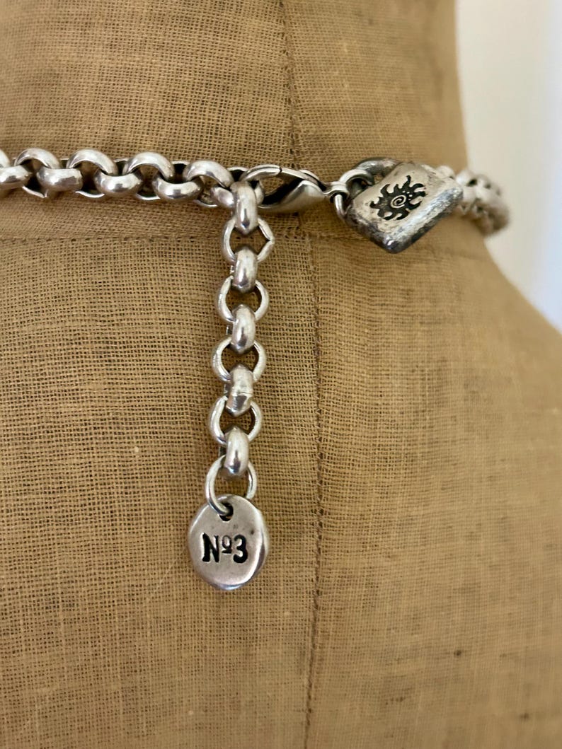 May include: Close-up of a silver-toned necklace with a large link chain. The necklace has a decorative clasp with a sunburst design and a small circular charm with the number "N&ordm;3" engraved on it. The necklace is displayed on a tan fabric.