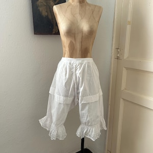 May include: White cotton bloomers featuring a high-waisted design with a drawstring closure at the center front. The garment includes a horizontal tiered seam above the knee and is finished with delicate white eyelet lace trim at the leg openings.