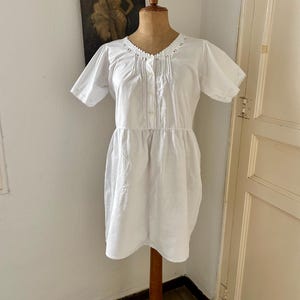 May include: A white short-sleeved dress featuring a round neckline with delicate pleating and button detailing down the center front. The garment has a gathered waistline and scalloped edges on the sleeves, presented on a wooden mannequin torso.