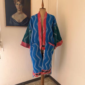 Peut inclure: Une robe à motifs bleus, rouges et verts, présentée sur un mannequin en bois. La robe présente un motif géométrique avec des coutures blanches. La robe est ouverte, révélant un intérieur et une bordure rouges. Un portrait encadré est en arrière-plan.