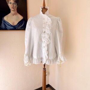 May include: A white blouse with a high ruffled collar and long sleeves, displayed on a wooden dress form. The blouse features a row of buttons down the front and delicate lace trim. The sleeves have lace cuffs with yellow accents.
