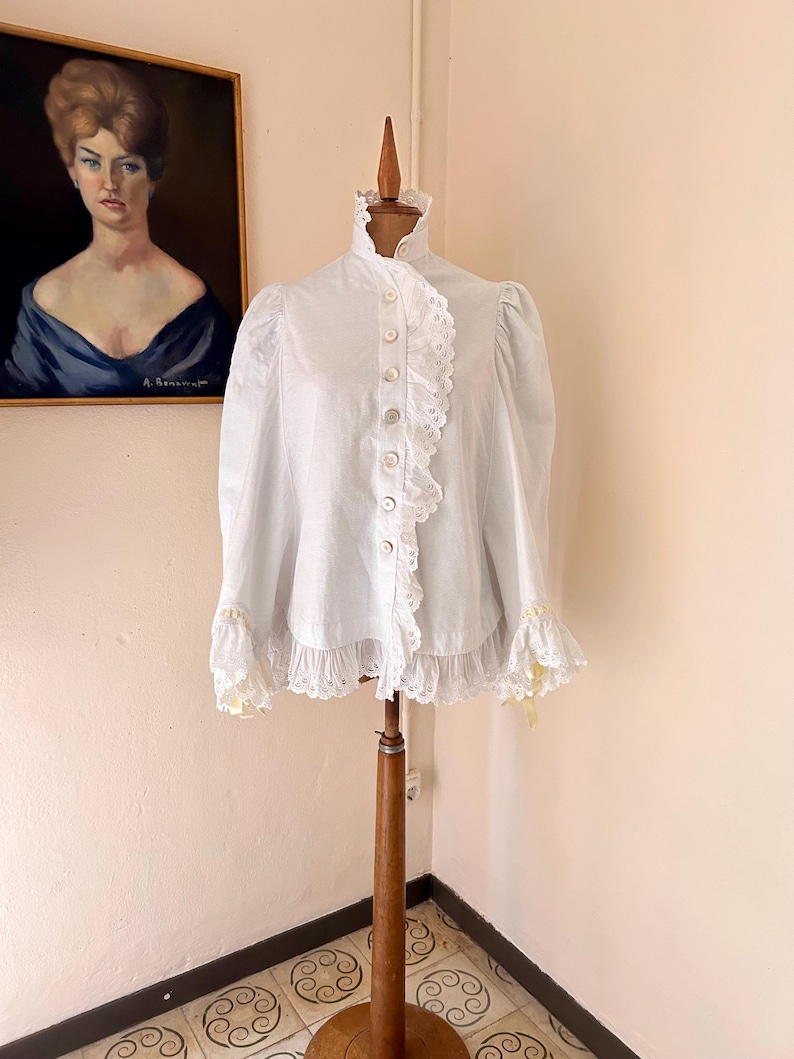 May include: A white blouse with a high ruffled collar and long sleeves, displayed on a wooden dress form. The blouse features a row of buttons down the front and delicate lace trim. The sleeves have lace cuffs with yellow accents.