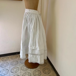 Puede incluir: Falda blanca de estilo vintage, con cintura fruncida y dobladillo con volantes. La falda se exhibe en un maniquí y presenta delicados adornos de encaje en el borde inferior. La tela parece ligera y fluida.