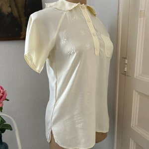 May include: A white, short-sleeved blouse with a collared neckline and a button-up front. The blouse has a small pocket on the left side and is decorated with delicate embroidered flowers.