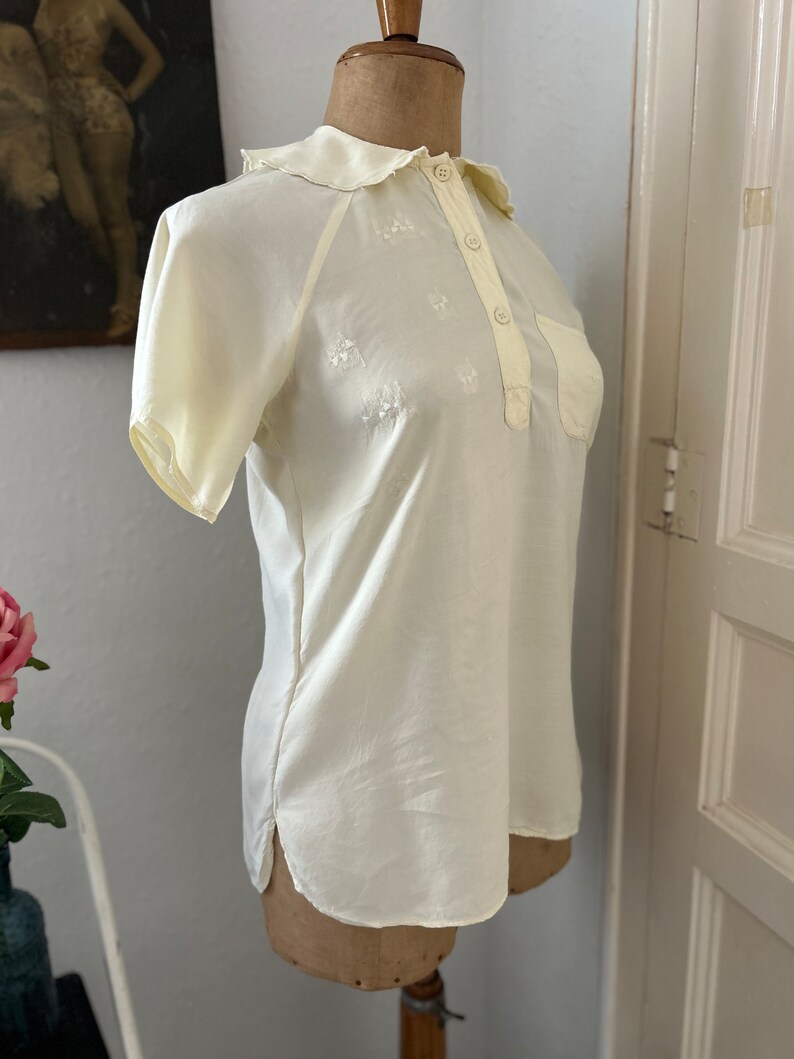 May include: A white, short-sleeved blouse with a collared neckline and a button-up front. The blouse has a small pocket on the left side and is decorated with delicate embroidered flowers.