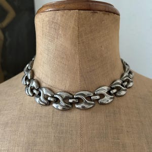 Puede incluir: Un collar de cadena de metal plateado que presenta una serie de eslabones interconectados, lisos y curvos. El collar tiene un acabado pulido y reflectante, mostrando un diseño uniforme de segmentos redondeados, sólidos y repetitivos.