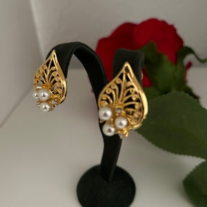May include: A pair of gold-tone earrings with a leaf design and pearl accents. The earrings are displayed on a black stand.