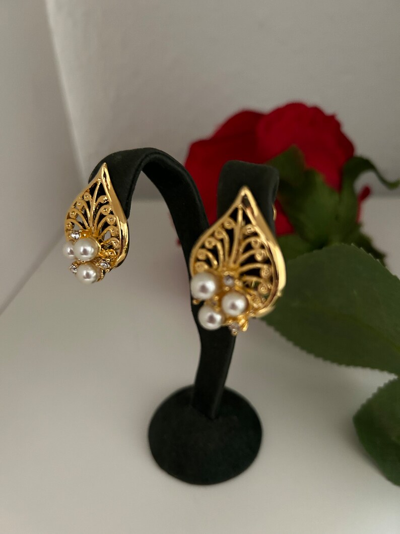 May include: A pair of gold-tone earrings with a leaf design and pearl accents. The earrings are displayed on a black stand.