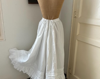 May include: A long white cotton skirt featuring a drawstring waist, vertical pintuck pleats near the hem, and a wide ruffled border with decorative cutwork detailing. The skirt is displayed on a mannequin, showing a full, flowing silhouette.
