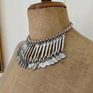 May include: Silver-toned statement necklace with a chain and a series of vertical bars and small, round pendants. The necklace is displayed on a vintage mannequin, showcasing its design and texture.