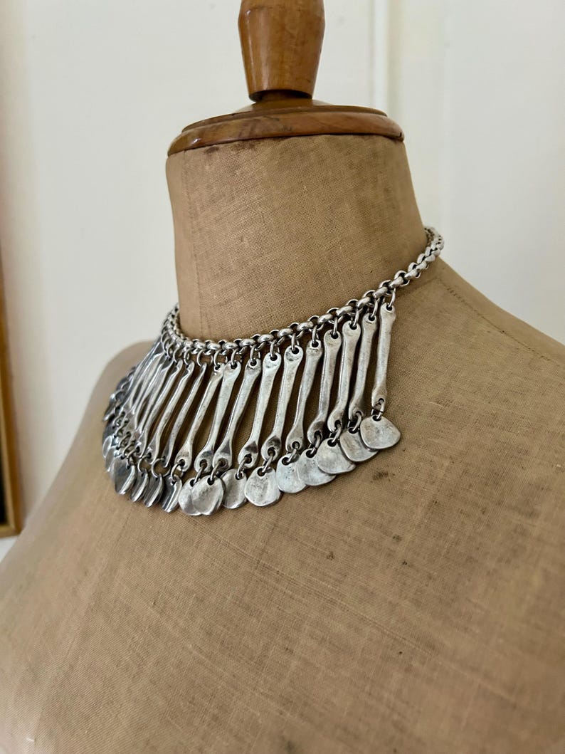 May include: Silver-toned statement necklace with a chain and a series of vertical bars and small, round pendants. The necklace is displayed on a vintage mannequin, showcasing its design and texture.