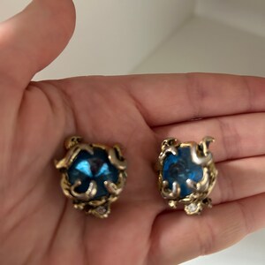 K&ouml;nnte beinhalten: Ein Paar Ohrringe im Vintage-Stil. Jeder Ohrring hat einen gro&szlig;en, facettierten blauen Edelstein, der in einen verzierten, goldfarbenen Metallrahmen gefasst ist. Der Rahmen hat ein wirbelndes Design mit kleinen, klaren Akzentsteinen.