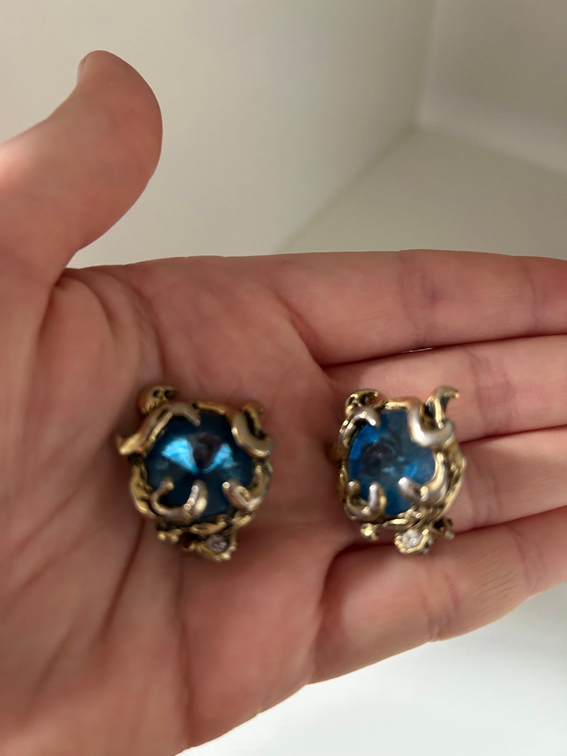 K&ouml;nnte beinhalten: Ein Paar Ohrringe im Vintage-Stil. Jeder Ohrring hat einen gro&szlig;en, facettierten blauen Edelstein, der in einen verzierten, goldfarbenen Metallrahmen gefasst ist. Der Rahmen hat ein wirbelndes Design mit kleinen, klaren Akzentsteinen.