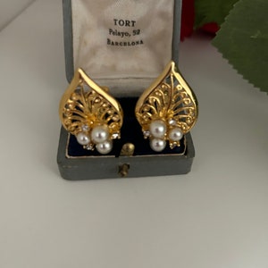 May include: A pair of gold tone clip-on earrings with a delicate filigree design and pearl accents. The earrings are in a blue velvet box with the inscription "TORT Pelayo, 52 BARCELONA".