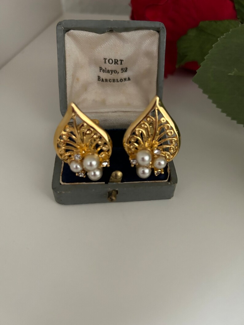 May include: A pair of gold tone clip-on earrings with a delicate filigree design and pearl accents. The earrings are in a blue velvet box with the inscription "TORT Pelayo, 52 BARCELONA".