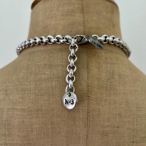 May include: A silver-toned necklace with a large link chain design. The necklace is fastened with a lobster clasp and has an adjustable chain. A small, round charm with the text "N93" hangs from the chain.