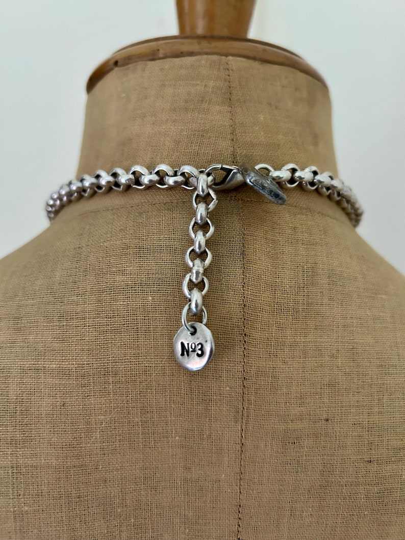 May include: A silver-toned necklace with a large link chain design. The necklace is fastened with a lobster clasp and has an adjustable chain. A small, round charm with the text "N93" hangs from the chain.