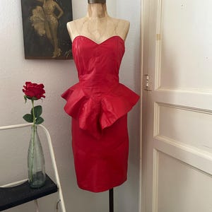 May include: A vibrant red, strapless dress with a peplum detail, displayed on a mannequin. The dress has a sweetheart neckline and a fitted silhouette. A vintage poster and a rose in a vase are in the background.