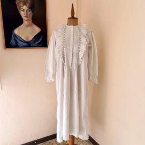 Op de afbeelding: Een witte vintage nachtjapon met lange mouwen en een hoge kraag met ruches. De nachtjapon wordt getoond op een houten paspop. De kraag en manchetten hebben delicate kanten details. Een ingelijst portret hangt op de achtergrond aan de muur.