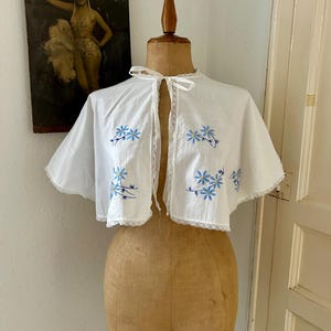 以下が含まれることがあります： 黄色い中心を持つ青い花の刺繍が施された白いケープレット。衣服の縁には繊細なレースのトリムがあしらわれ、ネックラインの中央には細い白いリボンが付いています。生地は軽量で、やや透け感のある外観をしています。