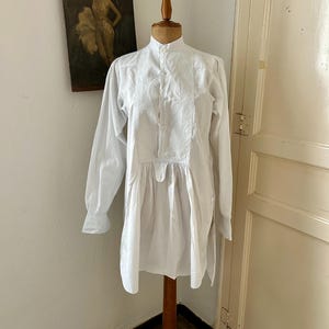 May include: A white long-sleeved shirt displayed on a wooden mannequin. The shirt features a high mandarin collar, a central button-down placket, and a pleated front panel that transitions into a loose, flowing hemline with side slits.