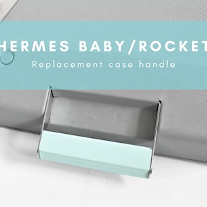 Pode incluir: Uma caixa de máquina de escrever cinza claro com uma alça azul e o texto "HERMES BABY/ROCKET Replacement case handle".