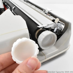 Pode incluir: Uma tampa de carretel de fita de máquina de escrever de plástico branco. A tampa é segurada na mão. A tampa é redonda com uma borda ligeiramente elevada.