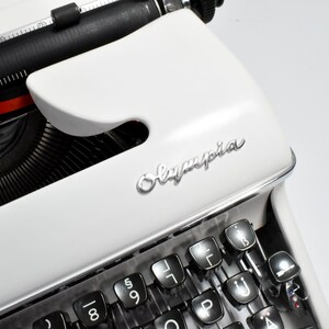 May include: Close-up of a white Olympia typewriter. The keyboard and keys are black, with the Olympia logo in silver. The typewriter's design is classic and vintage, with a focus on the keys and the brand name.