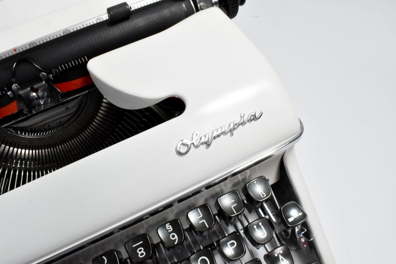 May include: Close-up of a white Olympia typewriter. The keyboard and keys are black, with the Olympia logo in silver. The typewriter's design is classic and vintage, with a focus on the keys and the brand name.
