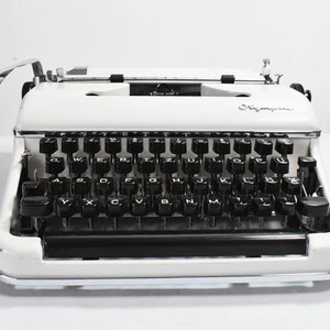 May include: A vintage white typewriter with black keys and a chrome carriage return lever. The typewriter has the word "Olympia" printed on the front. The keys are arranged in a QWERTY layout.