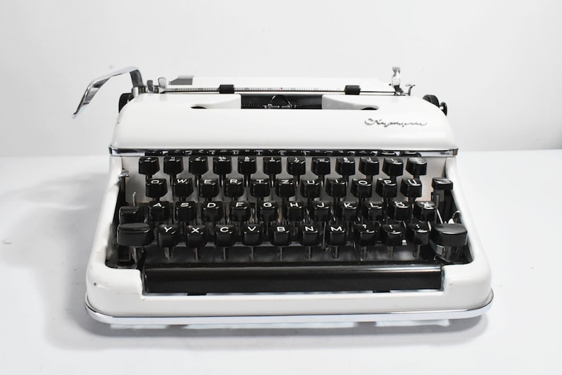 May include: A vintage white typewriter with black keys and a chrome carriage return lever. The typewriter has the word "Olympia" printed on the front. The keys are arranged in a QWERTY layout.