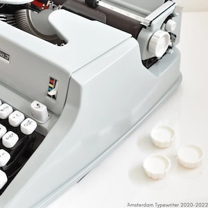 Pode incluir: Uma máquina de escrever Hermes Media 3 cinza com um teclado branco e teclas pretas. A máquina de escrever tem uma alavanca de retorno de carro branca e uma tecla de tabulação branca. Há três pés de plástico branco na mesa.