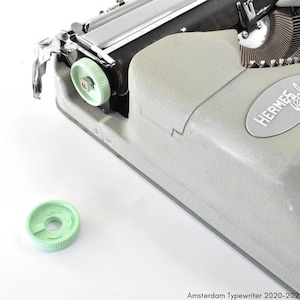 Pode incluir: Uma máquina de escrever Hermes Baby vintage verde com uma manivela de plástico verde e uma tampa de tecla de plástico verde. A máquina de escrever tem uma moldura de metal prateada e um teclado preto.