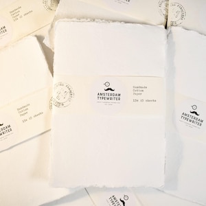 Puede incluir: Varios paquetes de papel de algodón blanco hecho a mano con bordes irregulares. Cada paquete lleva la etiqueta "AMSTERDAM TYPEWRITER" y "Handmade Cotton Paper 15x 15 sheets". El papel se presenta sobre una superficie de madera clara.