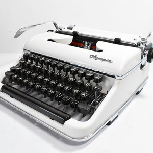 Puede incluir: Máquina de escribir Olympia vintage en blanco con teclas negras y detalles cromados. La máquina de escribir presenta un diseño clásico con una parte superior curvada y el logotipo de Olympia. Las teclas están dispuestas en un diseño QWERTY estándar.